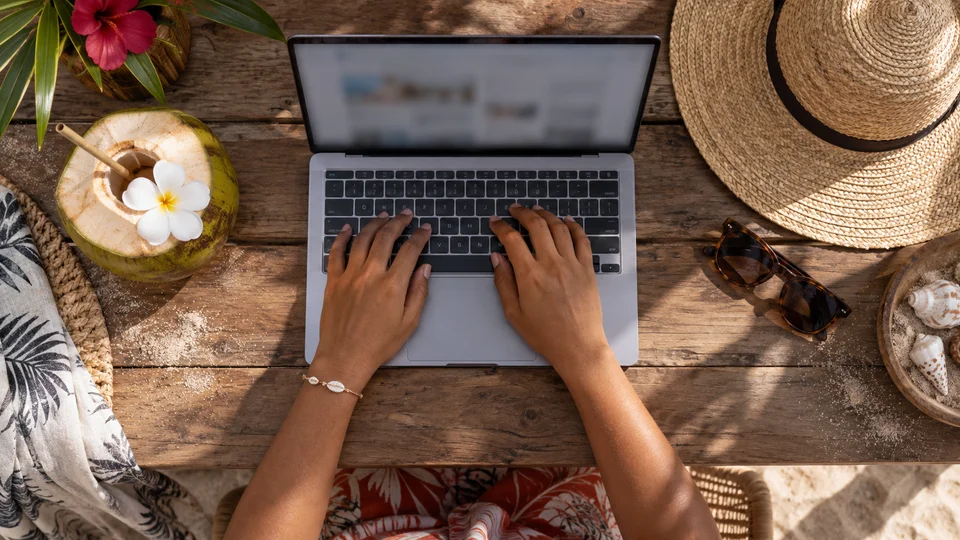 Digital nomad typing on a laptop at a wooden table with tropical beach accessories.