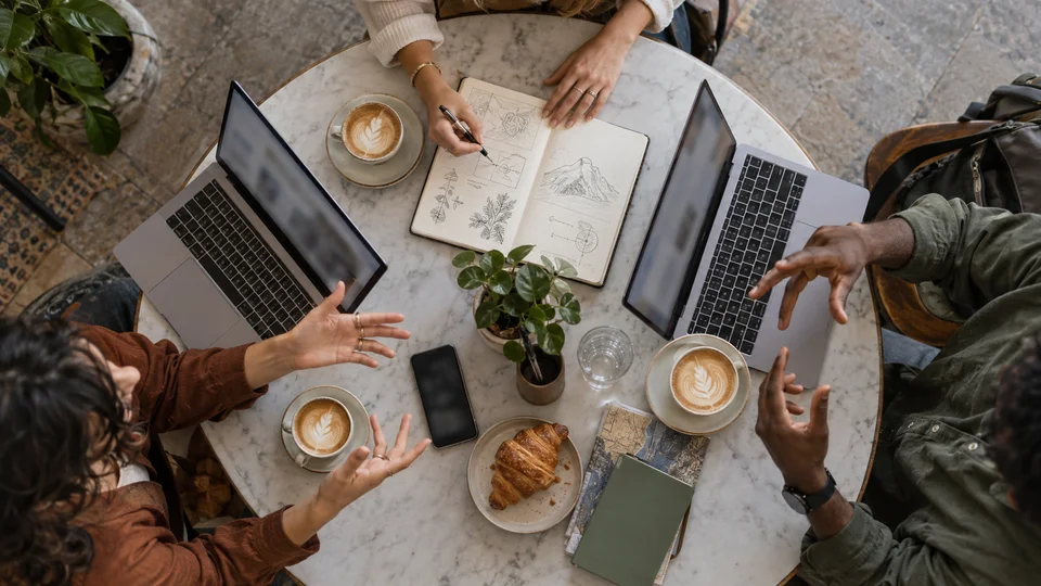 Two people talking at a café table with notebooks, laptops, and coffee cups.