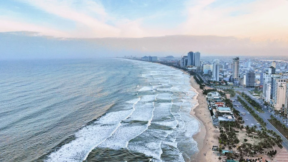 Sandy beach and seafront of a Vietnamese coastal city at dawn, with waves and high-rise buildings.