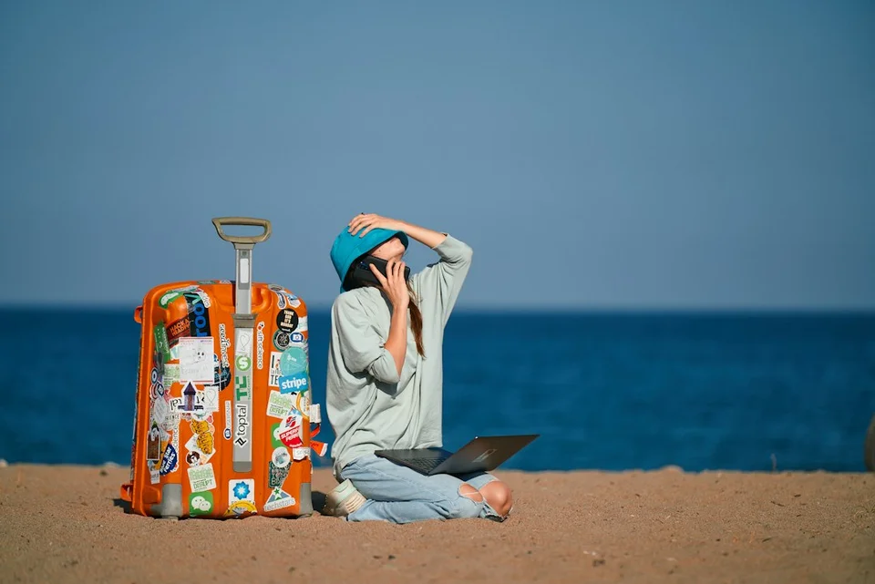 Digital nomad working on a laptop at the beach with a travel suitcase and the ocean in the background.