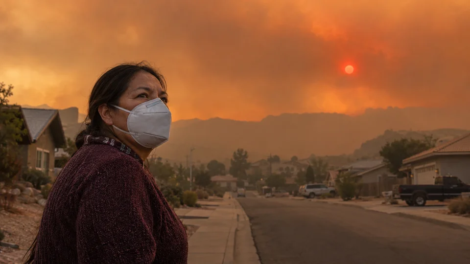 Woman wearing a mask facing wildfire smoke over hills at dusk in a residential area