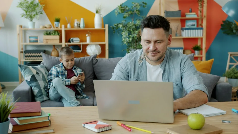 Parent working on a laptop at a home desk, with a child sitting on a nearby couch in a colorful, organized office.