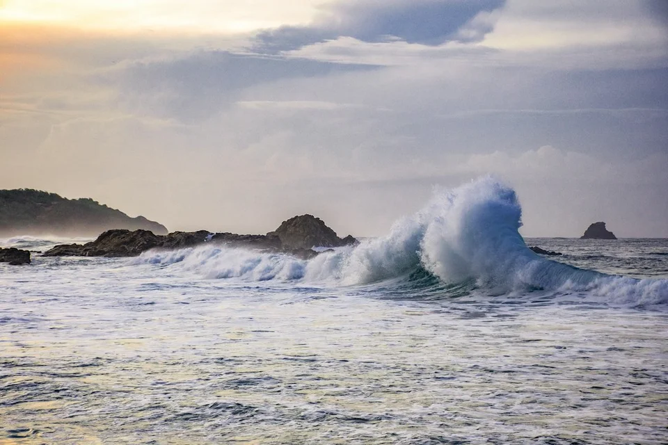 Huge waves crash against a rocky coastline under a stormy sky with dramatic clouds.