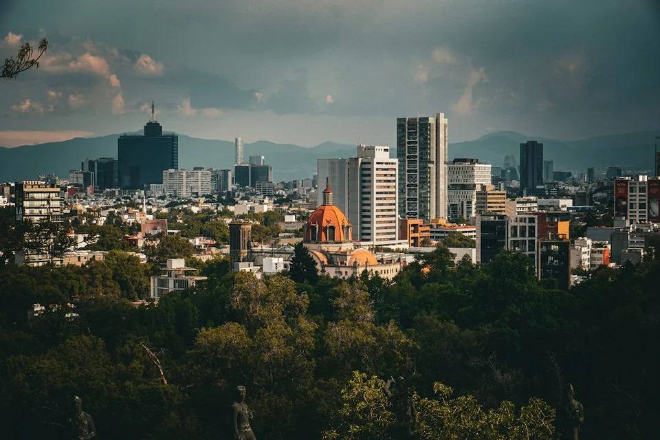Mexico City skyline at sunset with buildings, a church with a golden dome, and mountains in the background.