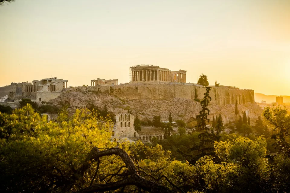 The Acropolis of Athens lit by the golden light of sunset, with olive trees and ancient ruins in the foreground.