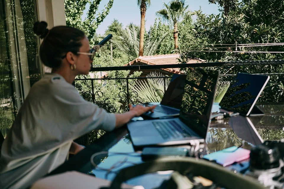 Digital nomad working on a laptop at an outdoor desk with tropical palm trees and greenery in the background.
